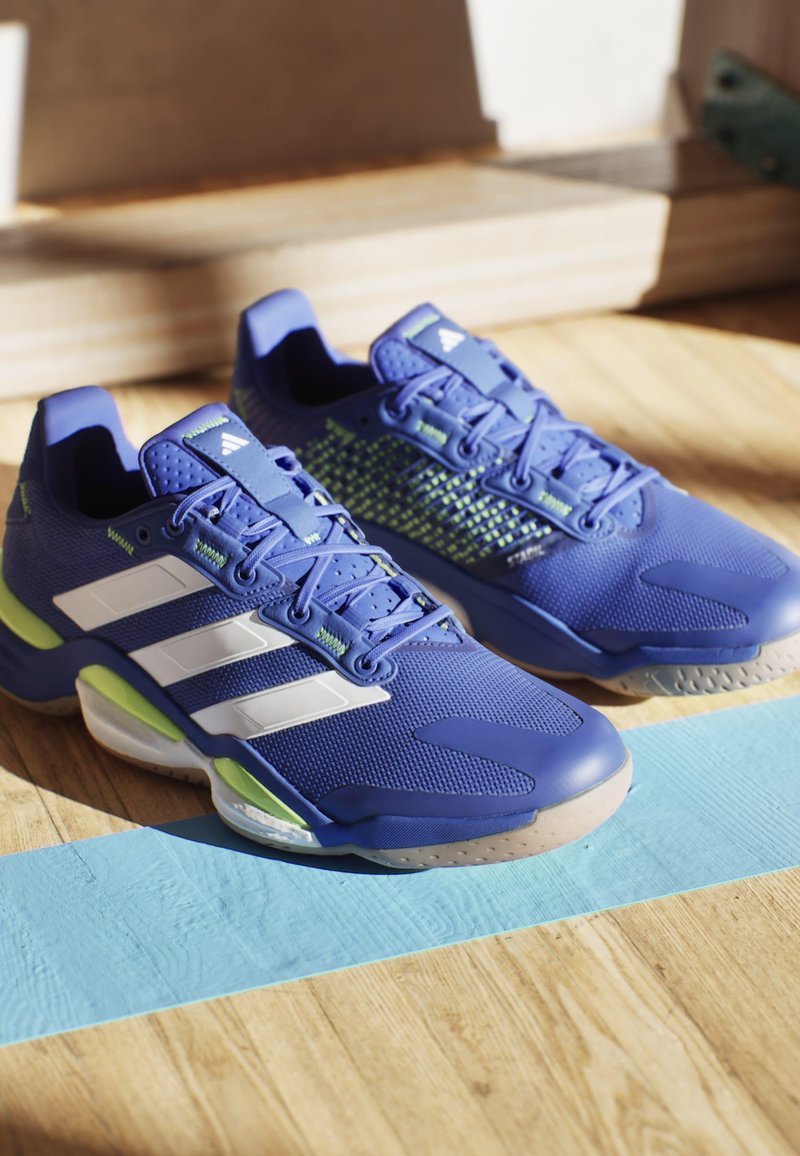 Blauwe sportschoenen van gaas en synthetisch materiaal. Voorzien van drie witte strepen, groene accenten, een textuurzool en vetersluiting.