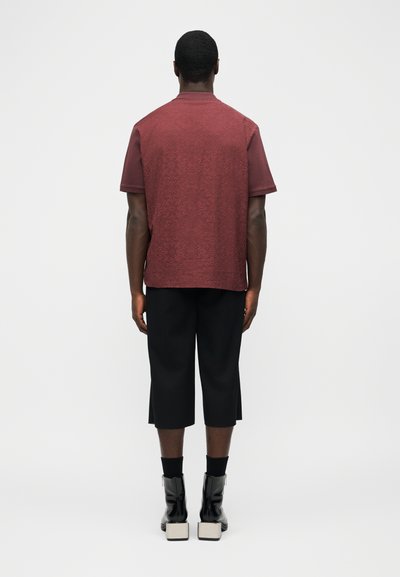 Άνδρας που στέκεται με την πλάτη στραμμένη φορώντας μακρυμάνικο πουκάμισο μπορντό, μαύρο παντελόνι μέχρι τον αστράγαλο, μαύρες κάλτσες και μαύρα μποτάκια με λευκές σόλες.