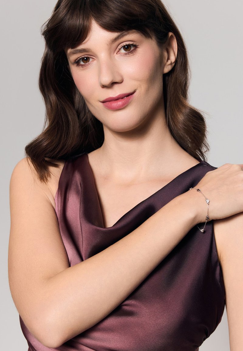 Srebrna bransoletka z serduszkowymi akcentami noszona na nagim ramieniu; modelka w głębokiej burgundowej satynowej sukni z opadającym dekoltem.