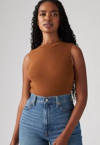 Brun sømløs topp laget av glatt, elastisk stoff, kombinert med høyderte lyseblå denimjeans. Teksturerte krøller rammer ansiktet.