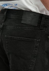 Czarne jeansy z denimu z skórzaną naszywką z tłoczonym tekstem. Posiadają tylnią kieszeń z ozdobnym, zakrzywionym przeszyciem.