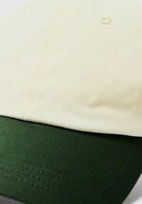 Keps med en krämfärgad ovansida och mörkgrön skärm, tillverkad av texturerad tyg; har synliga sömmar och en knapp på toppen.