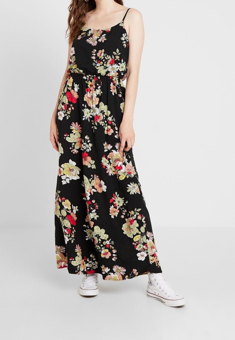 Maxiklänning i svart tyg, dekorerad med färgglada blommönster. Har tunna stammar och ett elastiskt midjeband, ihopparad med vita sneakers.