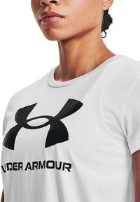 Camiseta blanca de manga corta con un gran logo negro de Under Armour y texto. La tela parece suave con un diseño clásico de cuello redondo.