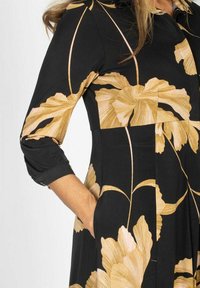 Czarna sukienka z złotymi wzorami kwiatowymi, długimi rękawami z podwiniętymi mankietami i kieszeniami po bokach. Uszyta z gładkiego materiału o dopasowanym kroju.