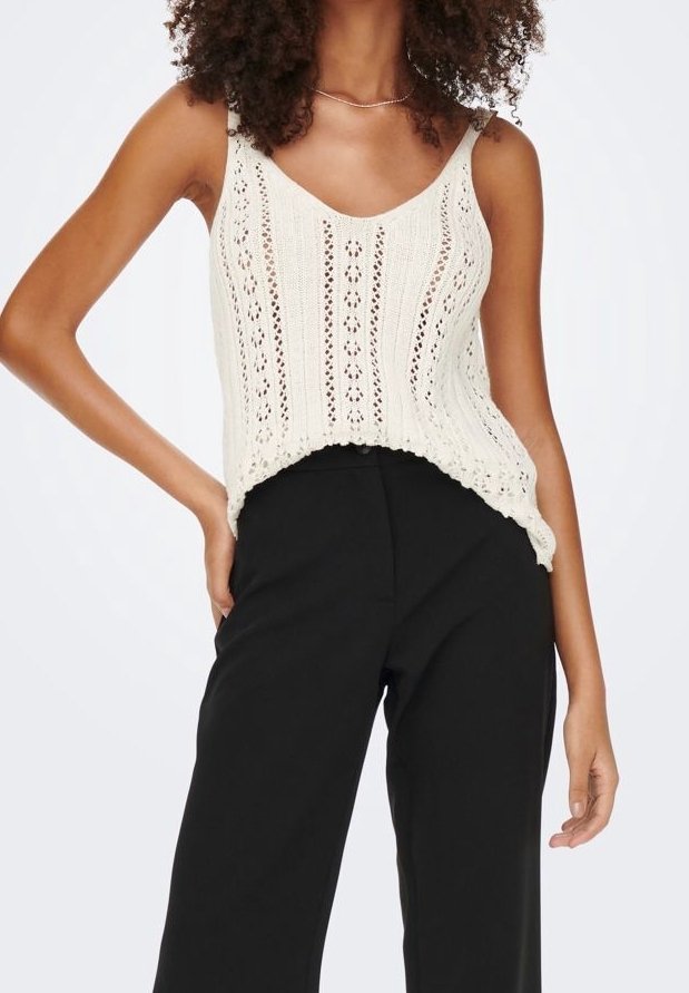 Femme portant un top sans manches en maille crème avec des motifs ajourés, associée à un pantalon large noir taille haute, posant avec la main sur la hanche.