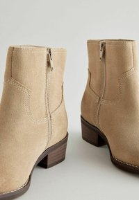 Stivali ankle in suede beige con cerniere laterali, dettagli cuciti e tacco in legno impilato. Texture liscia con cuciture rinforzate.