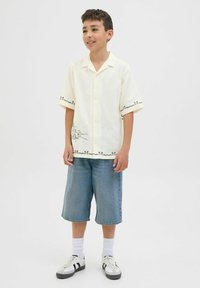 Kortärmad krämfärgad skjorta med svarta grafiska detaljer och accenter. Kombineras med ljusblå denimshorts och vita sneakers med svarta ränder.