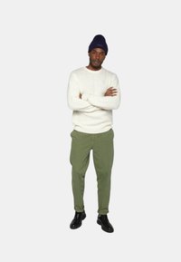 Pull en tricot blanc, pantalon vert olive et chaussures noires. Le modèle porte un bonnet bleu marine, bras croisés, se tenant debout sur un fond neutre.