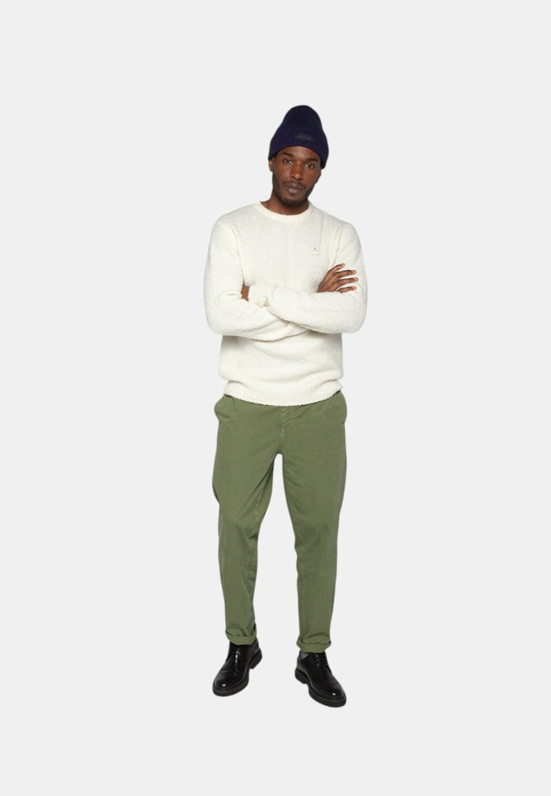 Pull en tricot blanc, pantalon vert olive et chaussures noires. Le modèle porte un bonnet bleu marine, bras croisés, se tenant debout sur un fond neutre.