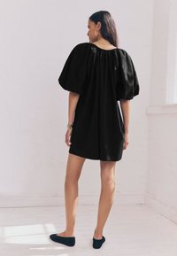 Sort kjole med pufærmer, løs pasform og rynket halsudskæring, lavet af letvægtsstof. Modellen har mørke sko og et ur på.
