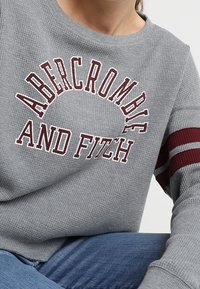 Szary, teksturowany sweatshirt z białym logo "ABERCROMBIE AND FITCH", z bordowymi paskami na rękawach. W połączeniu z niebieskimi dżinsami.