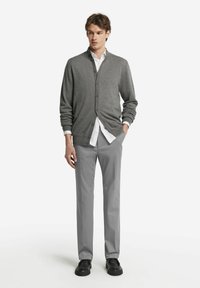 Grå cardigan med knappar, bärs över en vit skjorta. Kombineras med ljusgrå byxor och svarta skor. Enkelt och figursytt design.