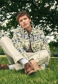 Junger Mann, der im Schneidersitz auf dem Gras unter einem Baum sitzt. Er trägt eine gemusterte Jacke, beigen Hosen, weiße Socken und braune Lederschuhe und wirkt nachdenklich.