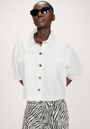 Personne portant des lunettes de soleil noires en forme d'œil de chat, une chemise blanche à manches courtes boutonnée et un pantalon imprimé zèbre noir et blanc sur un fond uni.