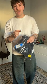 Μαύρο μπλουζάκι σε συσκευασία με μπλε ετικέτα κρατημένη από άτομο που φοράει λευκό φούτερ και φαρδιά πράσινα τζιν. Στο φόντο διακρίνονται μια τσάντα και διακοσμητικά.