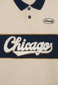 Camisa polo beige con cuello azul marino, dos botones y un logotipo bordado de "Chicago" en texto blanco en relieve, acentuado con un borde dorado.