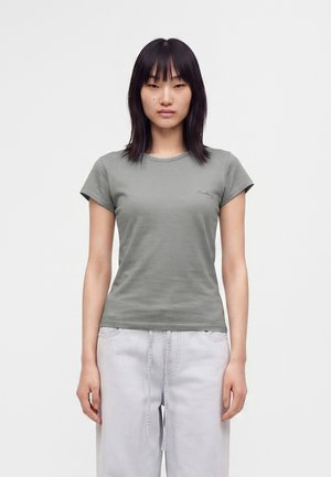 Jeune femme aux cheveux noirs et raides portant un t-shirt moulant à manches courtes vert olive et un pantalon gris clair sur un fond uni.
