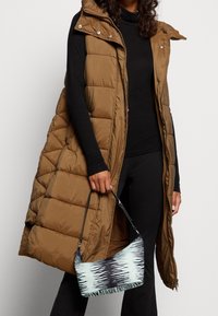 Kobieta w długiej brązowej puchowej kamizelce i czarnym stroju, trzymająca małą niebieską torbę na ramię w czarne wzory.