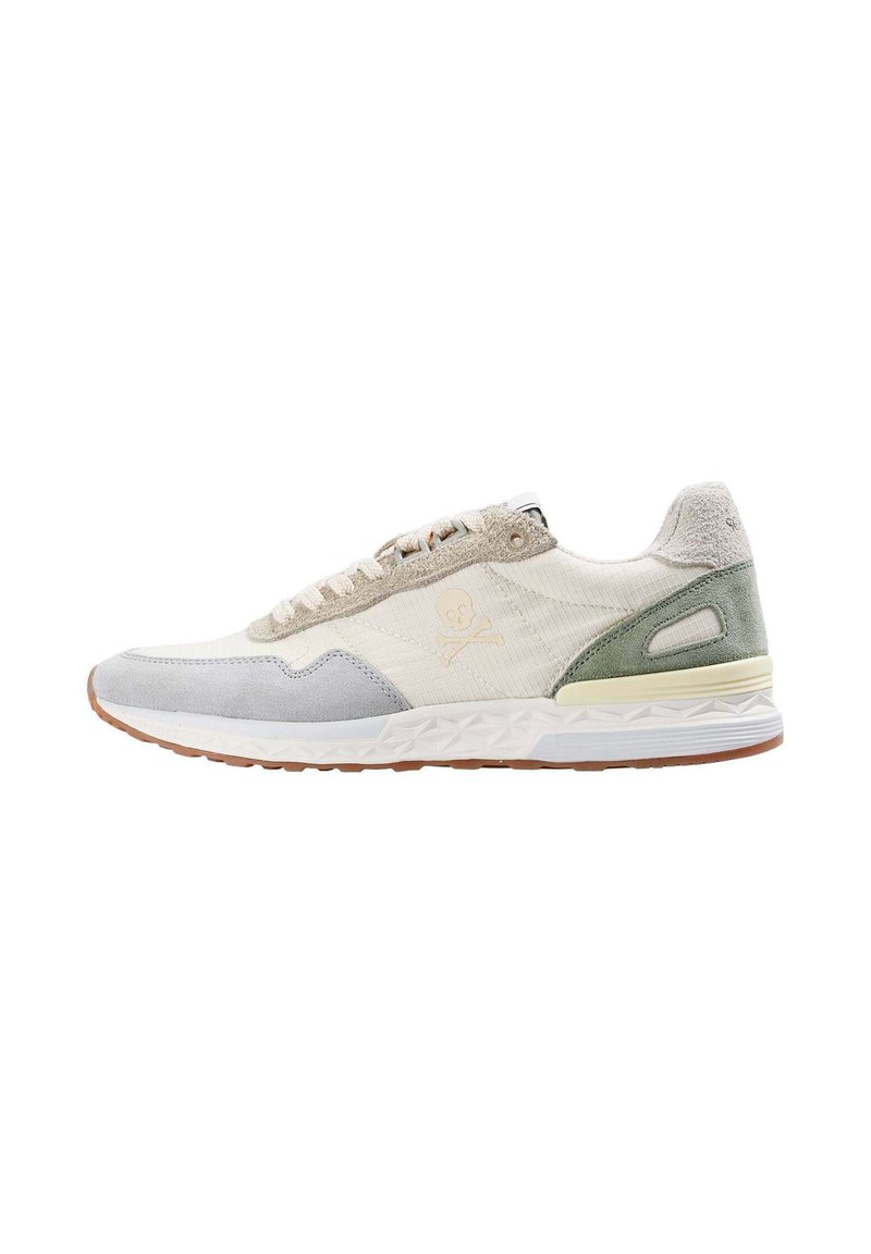 Zapatilla baja con acentos en ante beige, gris y verde, un logo de calavera y tibias en el lateral, y una suela blanca texturizada con una suela de goma.