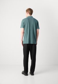 Kortermet, teal polo-skjorte laget av bomullsblanding, sammen med svarte, løstsittende bukser og svarte sko, sett bakfra.