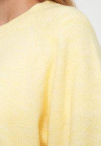 Dzeltens adīts džemperis ar apaļu kakla izgriezumu un mīkstu tekstūru. Tam ir smalkas krāsu variācijas, radot gaisīgu, jauktu izskatu.