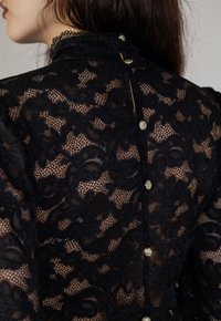 Schwarze Spitzenbluse mit hohem Kragen, floralem Muster und dekorativen goldenen Knöpfen entlang des Rückens. Transparente Textur und filigrane Details.