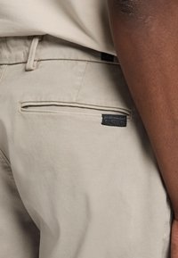 Beige broek met een gladde textuur, voorzien van een achterzak met een zwart label. Naaddetails zijn zichtbaar rond de zakken.