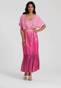 Rosa maxikjol med lös passform, V-ringning, korta ärmar, paljettextur och volangnederkant. Kombinerad med ljusa plattformssandaler.