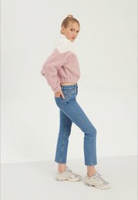 Šviesiai rožinis flisinis megztinis su balta apykakle, derinamas su mėlynais aukšto liemens džinsais ir smėlio spalvos sportiniais bateliais, parodytas šoniniu pozavimu.