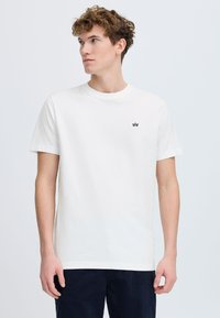 Hvit bomulls t-skjorte med rund hals og korte ermer. Har en liten svart logo på venstre brystområde. Minimalistisk design.