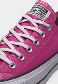 Ροζ αθλητικό παπούτσι Converse All Star χαμηλού ύψους με λευκά κορδόνια, μεταλλικές οπές, ορατή ραφή και λευκή ελαστική σόλα με μαύρη ρίγα.