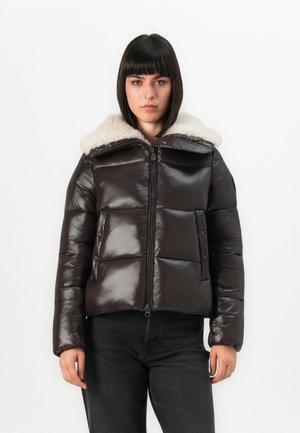 Vrouw met steil donker haar, gekleed in een glanzende zwarte pufferjack met een witte schapenvachtkraag, staande tegen een effen witte achtergrond.