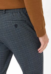Pantaloni a quadri da uomo in blu scuro con motivi grigi e senape, dotati di tasca posteriore con chiusura a bottone e un tessuto testurizzato.