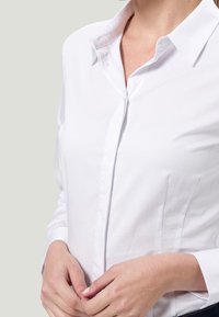 Camicia bianca con colletto, caratterizzata da una texture liscia e croccante e cuciture laterali. Chiusura frontale con bottoni e maniche lunghe in evidenza.