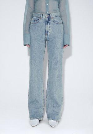 Osoba nosząca jasnoniebieskie dżinsy z wysokim stanem i szerokimi nogawkami, prześwitującą niebieską koszulę z długim rękawem oraz białe buty w szpic, stojąca na prostym tle.