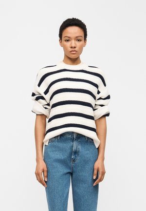 Personne portant un pull en maille blanche avec de larges rayures horizontales noires, un jean bleu taille haute, debout devant un fond clair uni.