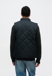 Σκούρο μπλε γιλέκο με καπιτονέ σχέδιο, ψηλό γιακά, ελαφριά επένδυση και χωρίς ορατά μεταλλικά στοιχεία, φορεμένο πάνω από σκούρο πουλόβερ και τζιν παντελόνι.