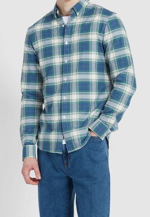 Hombre con camisa de cuadros verde, azul y blanca con botones, y vaqueros azules, de pie con una mano en el bolsillo.