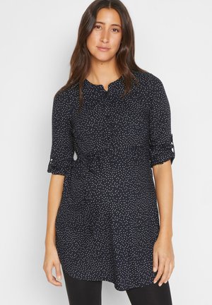 Tunic blu con pois bianchi, colletto abbottonato, maniche arrotolabili e vita elastica. Il tessuto morbido e la lunghezza leggermente maggiore offrono comfort.