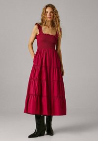 Naine seisab käed taskutes, kandes varrukateta punast kihtidest midi-kleid ja musti poolpikkade varbavahega kontsakingi ühetoonilise tausta ees.