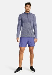 Blå långärmad träningströja med halvdragkedja, tillsammans med lila shorts. Texturerat tyg med en reflekterande logotyp på bröstet. Svarta sneakers.