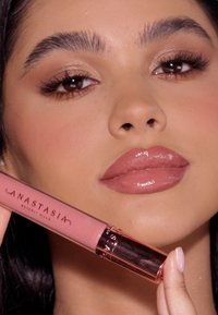 Vloeibare lippenstift in een roze tube met een gouden accent, vastgehouden in een hand. Het model heeft glanzende lippen en neutrale make-up. Bruine ogen en donker haar.