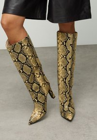 Knielange stiletto laarzen met een gestructureerd geel-zwart slangenprintontwerp, voorzien van een spitse neus en een nauwsluitend silhouet.
