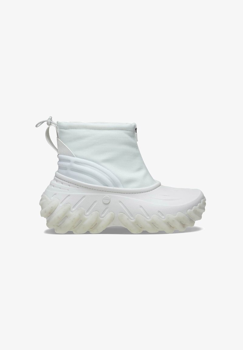 Zapatilla blanca con un alto collar de tobillo acolchado, material superior liso y una suela gruesa y texturizada con un patrón de olas. Incluye detalle de cremallera.