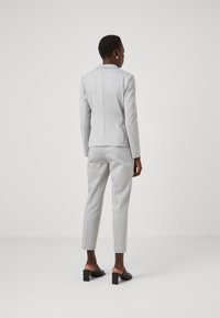 Ženska, oblečena v svetlo sivo prilegajočo se blazer in usklajene taperirane hlače, obrnjena stran od kamere, dodatki so črne pete mule in srebrni uhani.