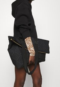 Czarna pikowana torba typu tote z geometrycznym wzorem, z złotym zamkiem błyskawicznym i dwoma solidnymi uchwytami. Tkanina ma teksturowane wykończenie.