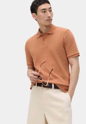 Mężczyzna w pomarańczowej polo z fakturą i kremowych spodniach, trzyma okulary przeciwsłoneczne w jednej ręce, patrząc w lewo.