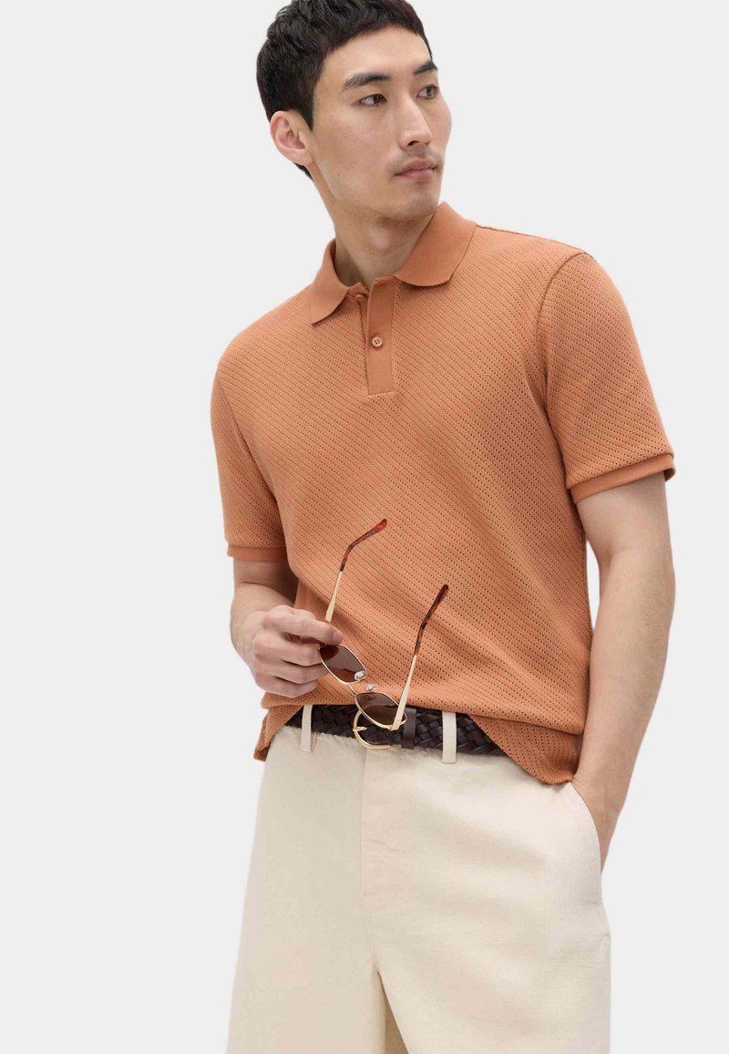 Mand iført orange tekstureret poloshirt og cremefarvede bukser, holder solbriller i den ene hånd og kigger til venstre.