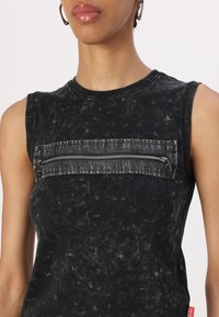 Svart ärmlös topp med en texturerad yta, med en horisontell ficka med dragkedja på framsidan och en liten röd etikett på sidan.